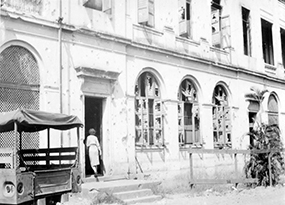 A black and white photo of the façade of De La Salle College, riddled with bullet holes, smashed windows and a military jeep parked near a doorway that a woman is walking in to.